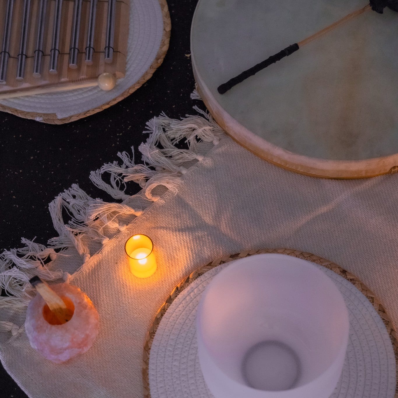 A crystal singing bowl, a frame drum, chimes and a lit candle arranged on a fringed cream throw