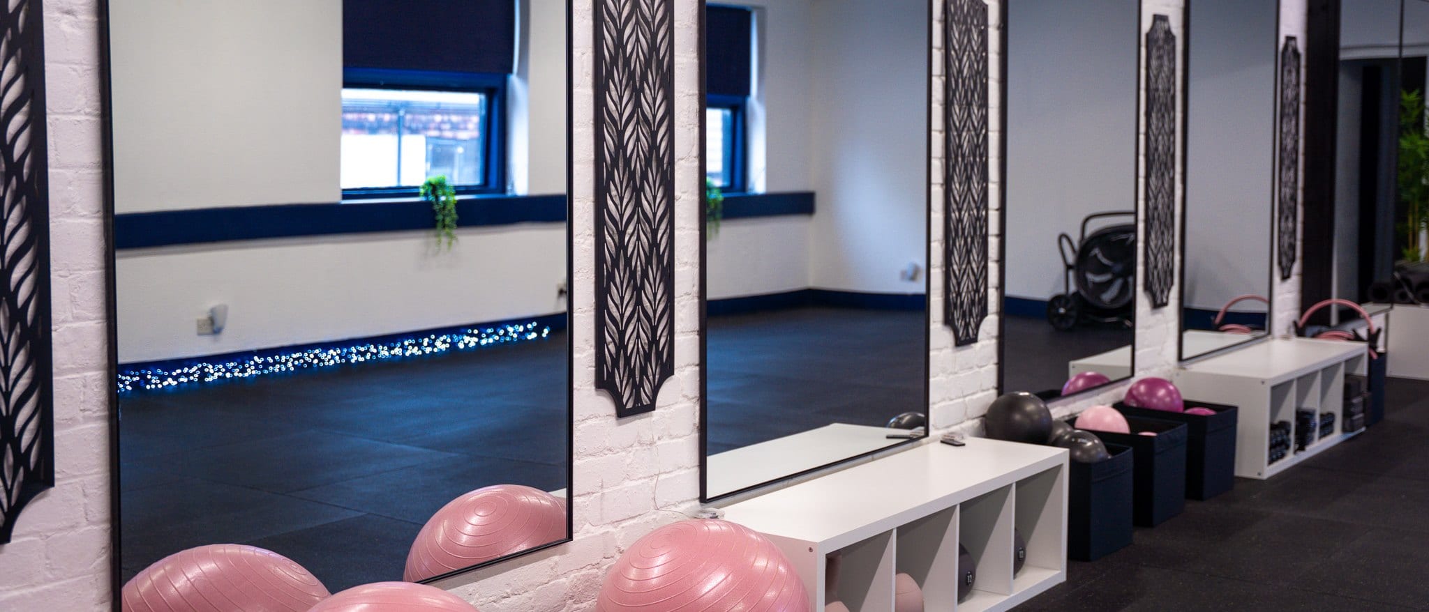 A wide view along the studio's mirrored wall, with pink stability balls and white storage benches beneath it