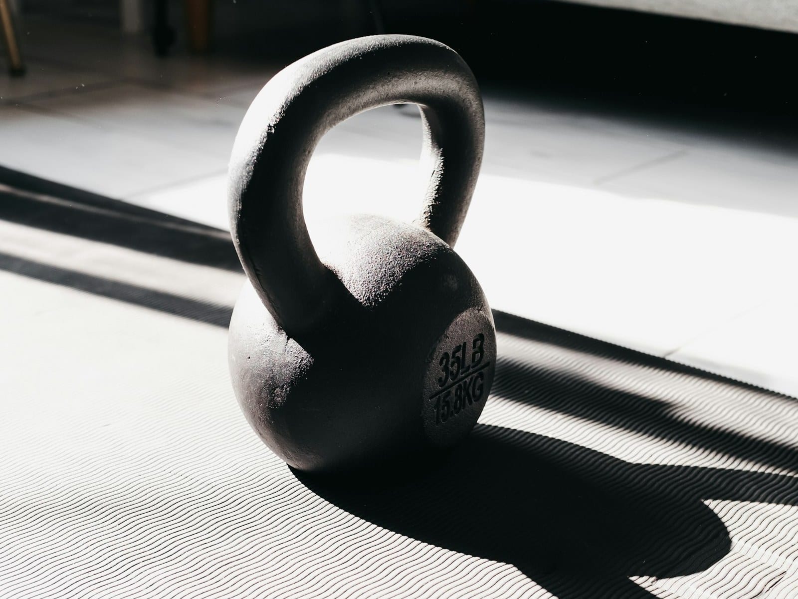 A single cast-iron kettlebell resting on a ribbed exercise mat in hard diagonal light