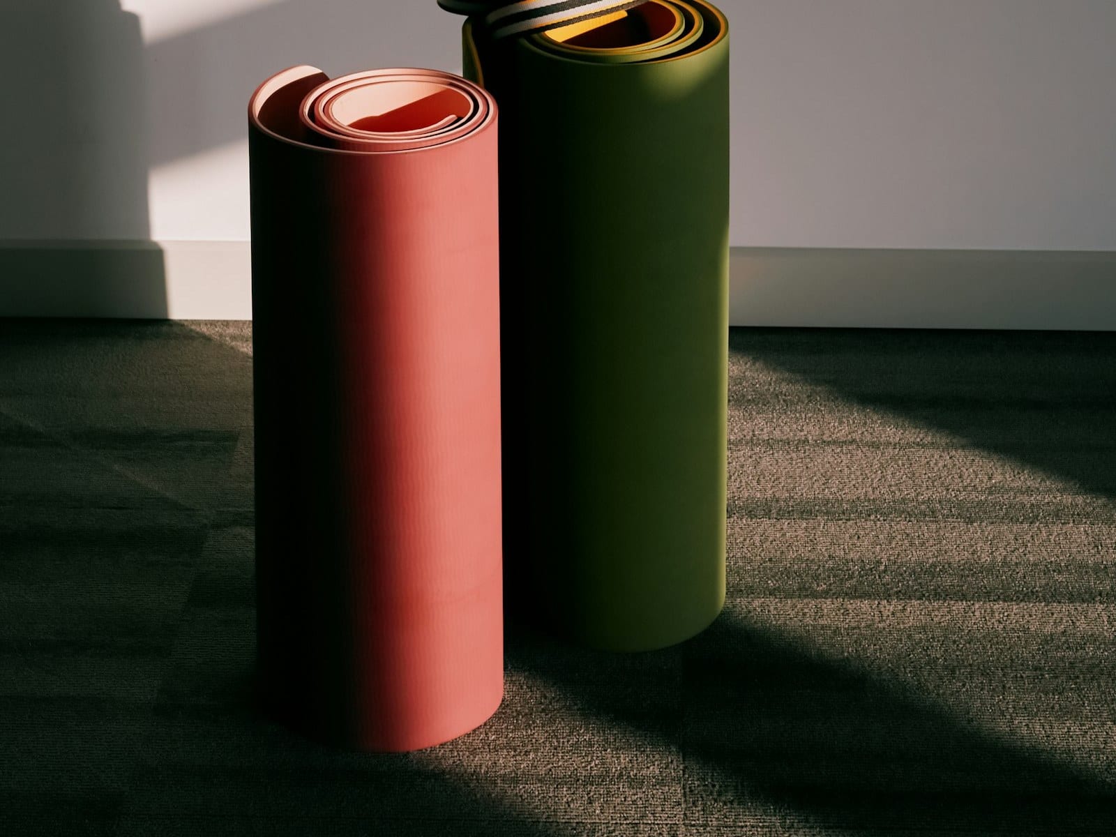 Two rolled exercise mats standing against a wall, one coral pink and one olive green, with a striped carrying strap looped over the top