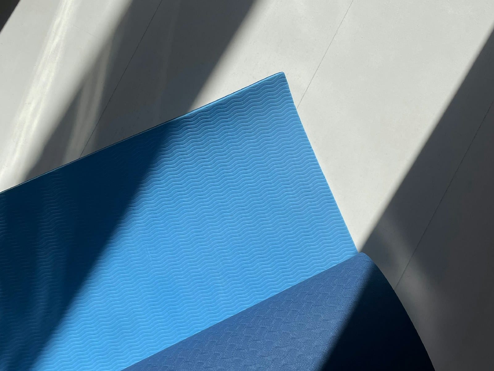 The corner of a two-tone blue exercise mat part-unrolled on a pale floor, lit by hard diagonal light